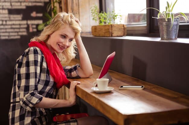 A young woman smiling and working from her laptop in a cozy coffee shop. She is dressed casually and appears relaxed, enjoying a beverage while she works. The background includes other people working and chatting, creating a vibrant yet laid-back atmosphere.
